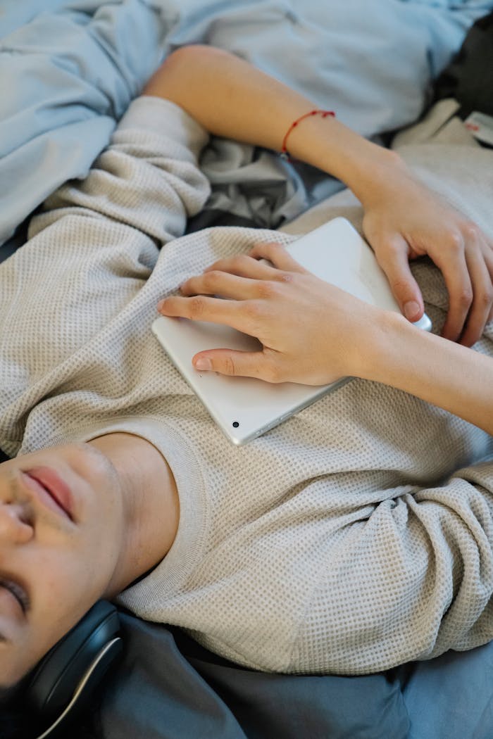 From above of crop ethnic male with tablet and closed eyes listening to music from headphones on bed at home