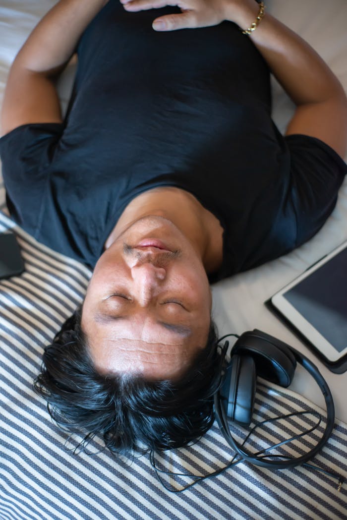 Man lying on bed with eyes closed, headphones, and tablet nearby, conveying relaxation.